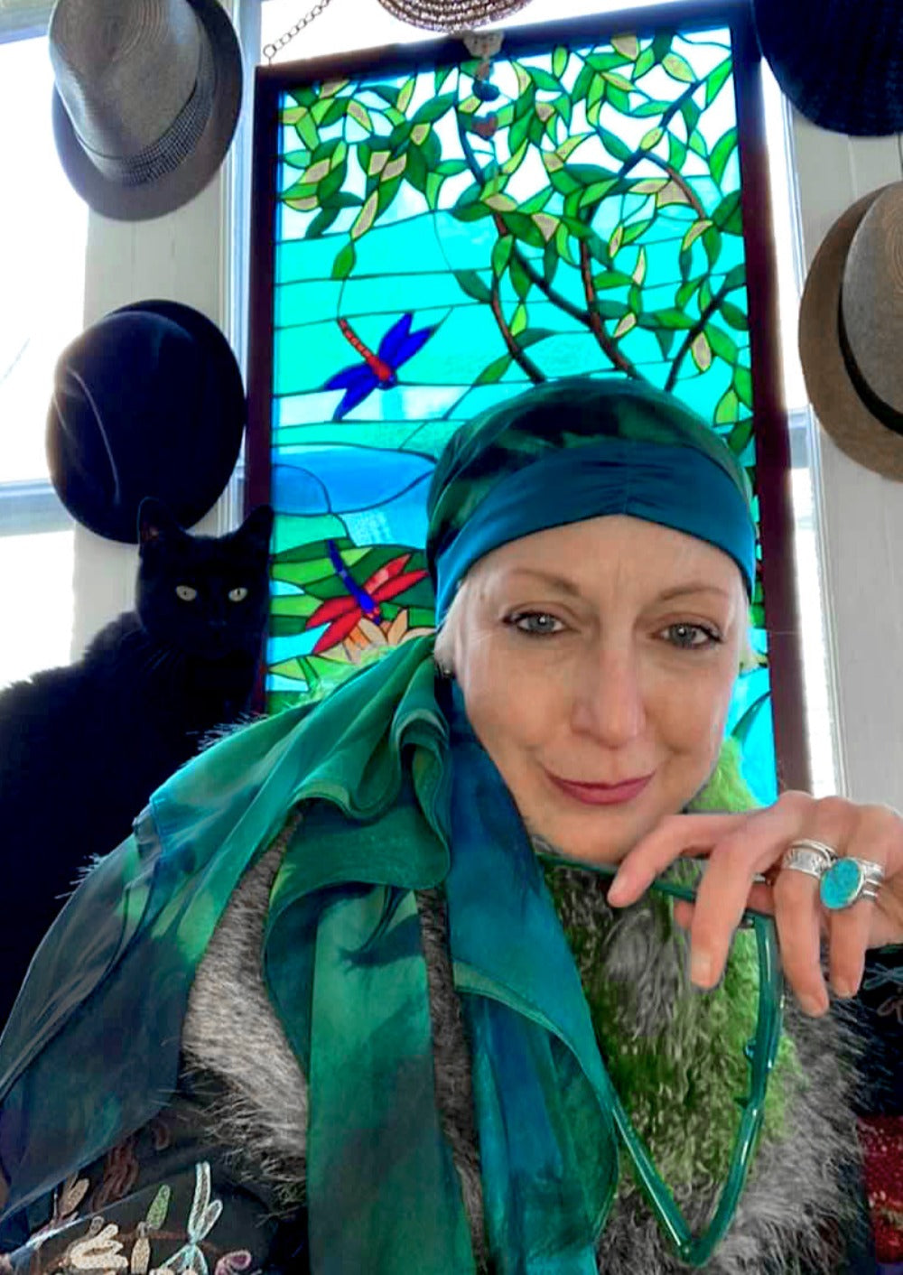 Bohemian artist woman, wearing a silk headscarf and a big turquoise ring, holding sunglasses and smiling. sitting in front of a stained glass window with a black cat at her side