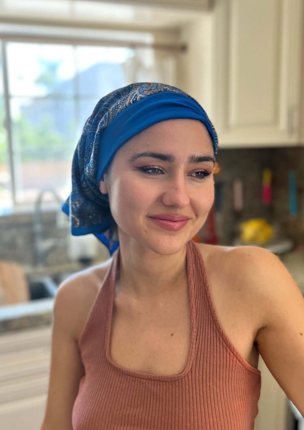 A beautiful lady in a jewel tone tank top, stands in her kitchen smiling. On her head she wears A paisley pattern of Deep blue with hints of golden, on a custom printed scarf, which is Made into a one piece headscarf with an organic cotton base in the sam