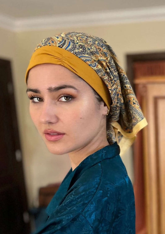 A beautiful lady in a deep blue silk pajama top, looks knowingly to the camera. On her head she wears A paisley pattern of golden with hints of blue, on a custom printed scarf, which is Made into a one piece headscarf with an organic cotton base in the sa