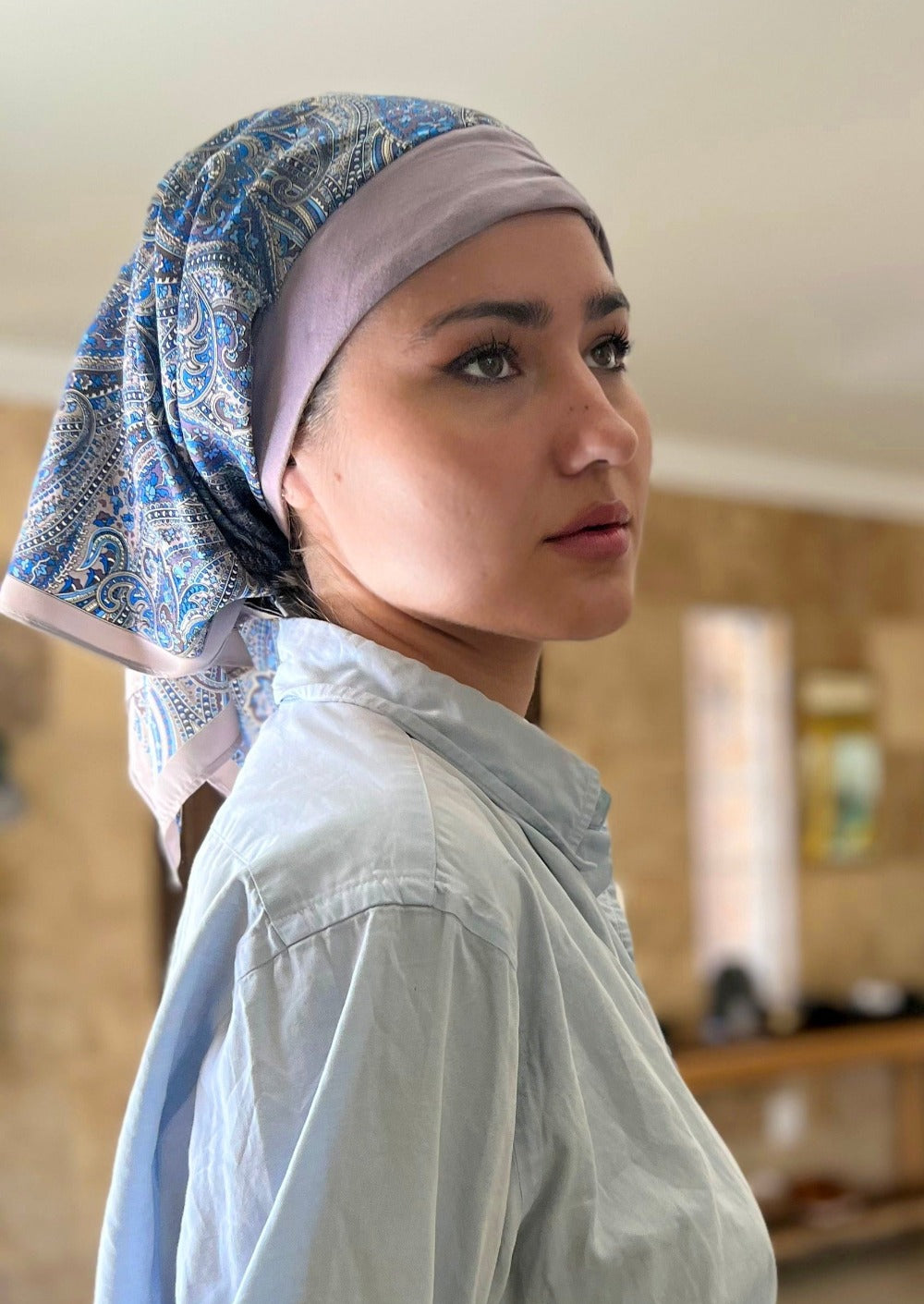 A beautiful woman, wearing a light jean shirt stand smiling in her kitchen. She wears a headscarf on her head, with A paisley pattern of lavender with hints of blue, on a custom printed scarf, It has an organic cotton base in the same calming lavender col