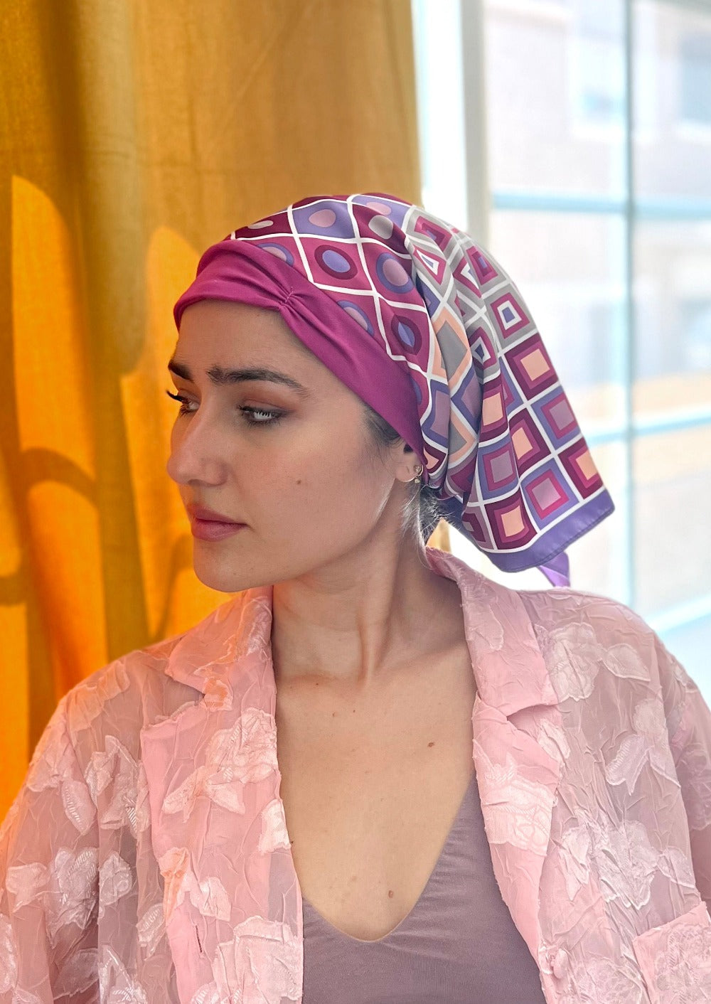 A beautiful woman gazes knowingly out the window, she has on a pink button down pj top. Her head is covered with a very attractive headscarf. The headscarf is made up of multipal layers, the base layer is a rich pink stretch cotton, that appears like a 