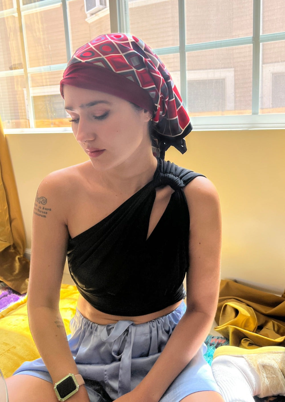 A beautiful woman, wearing an asymmetrical black shirt, sits looking down in her living room. She wears a headscarf on her head, the headscarf is made up of multiple layers, the base layer is a rich ruby stretch cotton, that appears like a headband, with 
