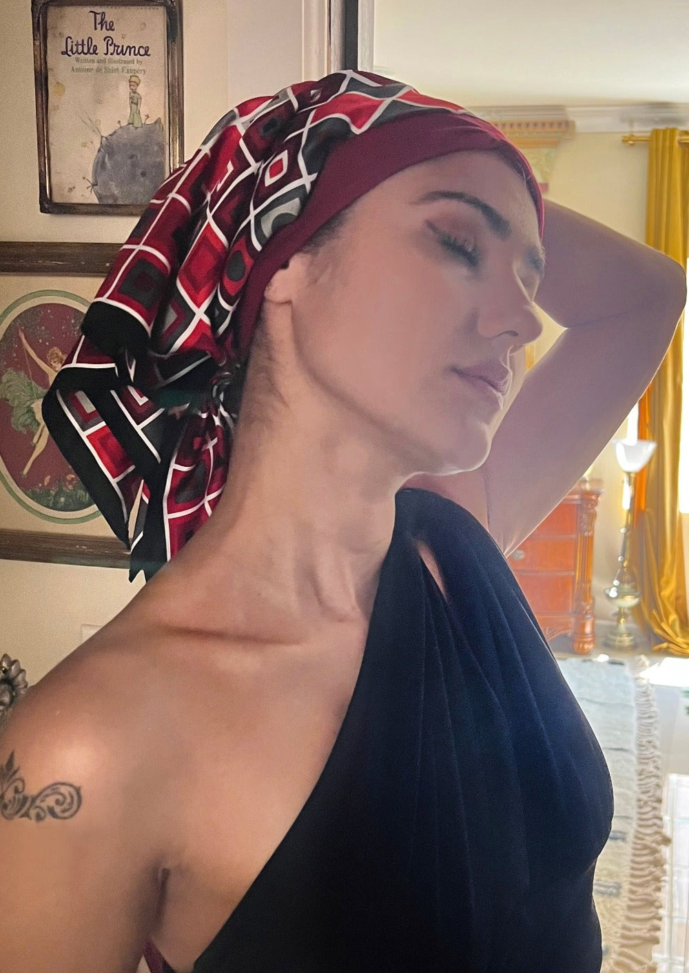 A beautiful woman, wearing an asymmetrical black shirt, stands looking to the side, in her living room. She wears a headscarf on her head, the headscarf is made up of multiple layers, the base layer is a rich ruby stretch cotton, that appears like a head