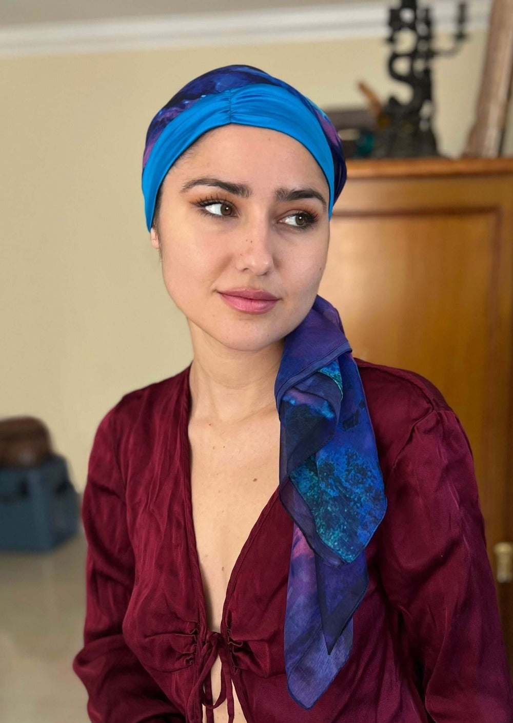 A woman wearing a bright blue silk & chiffon headscarf, smiles knowingly. She has a boysenberry colored long sleeve silk blouse on.