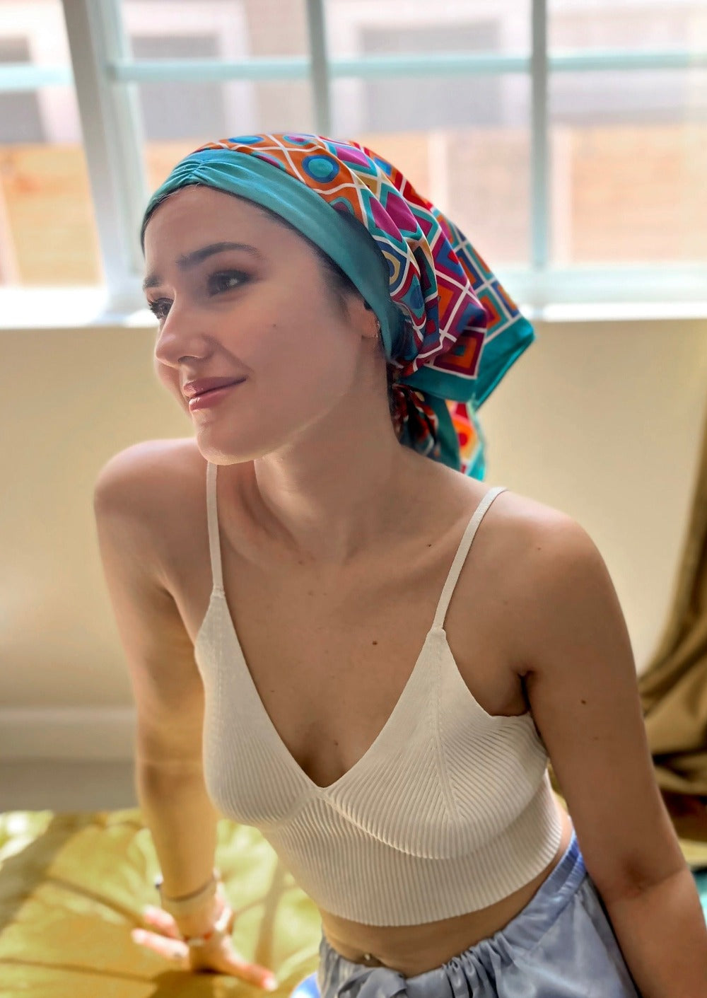 A beautiful woman, wearing an asymmetrical black shirt, sits smiling looking to the side, in her living room. She wears a headscarf on her head, the headscarf is made up of multiple layers, the base layer is a rich teal color stretch cotton, that appears