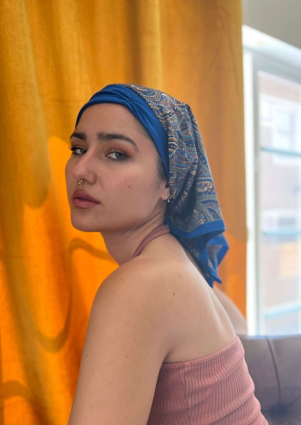 A beautiful lady in a jewel tone tank top, looks knowingly to the camera. On her head she wears A paisley pattern of Deep blue with hints of golden, on a custom printed scarf, which is Made into a one piece headscarf with an organic cotton base in the sa