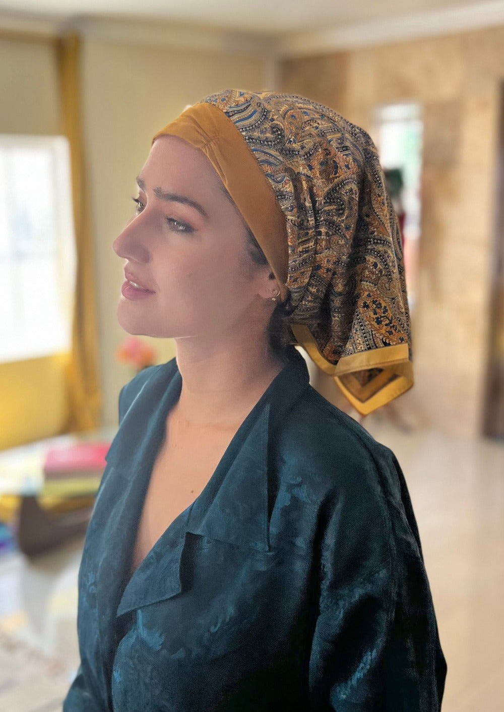 A beautiful lady in a deep blue silk pajama top, looks away from the camera, but is smiling. On her head she wears A paisley pattern of golden with hints of blue, on a custom printed scarf, which is Made into a one piece headscarf with an organic cotton b