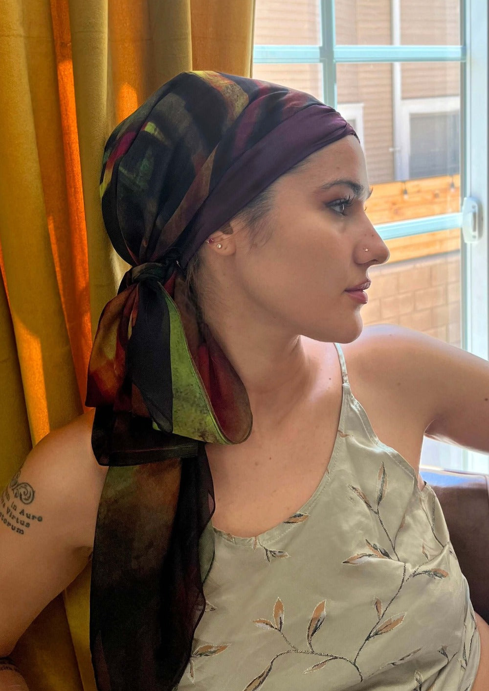 A beautiful woman, looking out a window shows off her luxurious chiffon and silk headscarf. It is an abstract print is wine colored with brush strokes of black, blood red and hints of bright green and chartreuse.