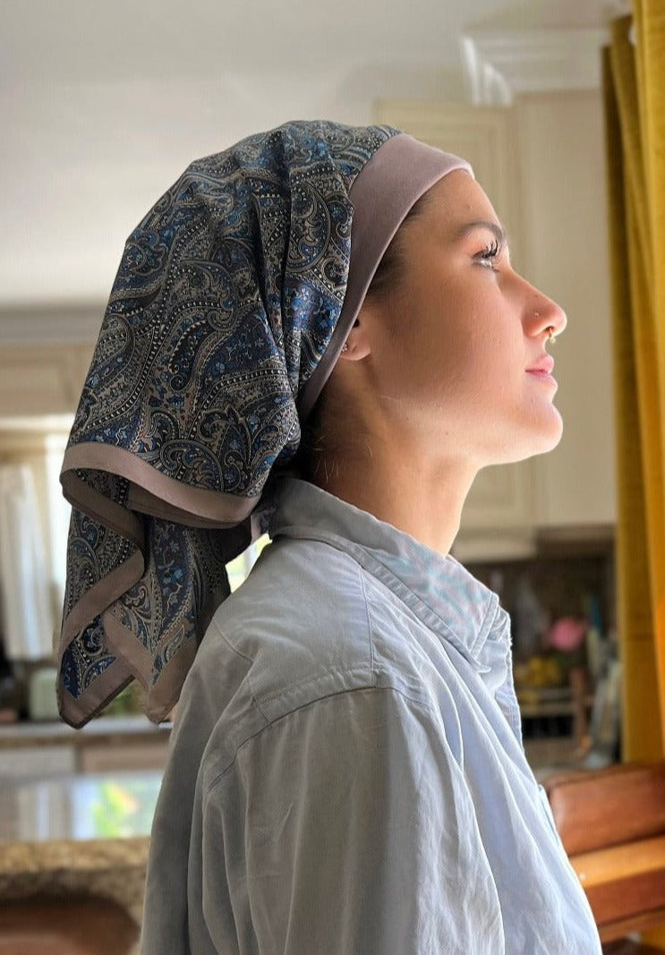 A beautiful woman, wearing a light jean shirt stands looking out the window in her kitchen. She wears a headscarf on her head, with A paisley pattern of lavender with hints of blue, on a custom printed scarf, It has an organic cotton base in the same calm