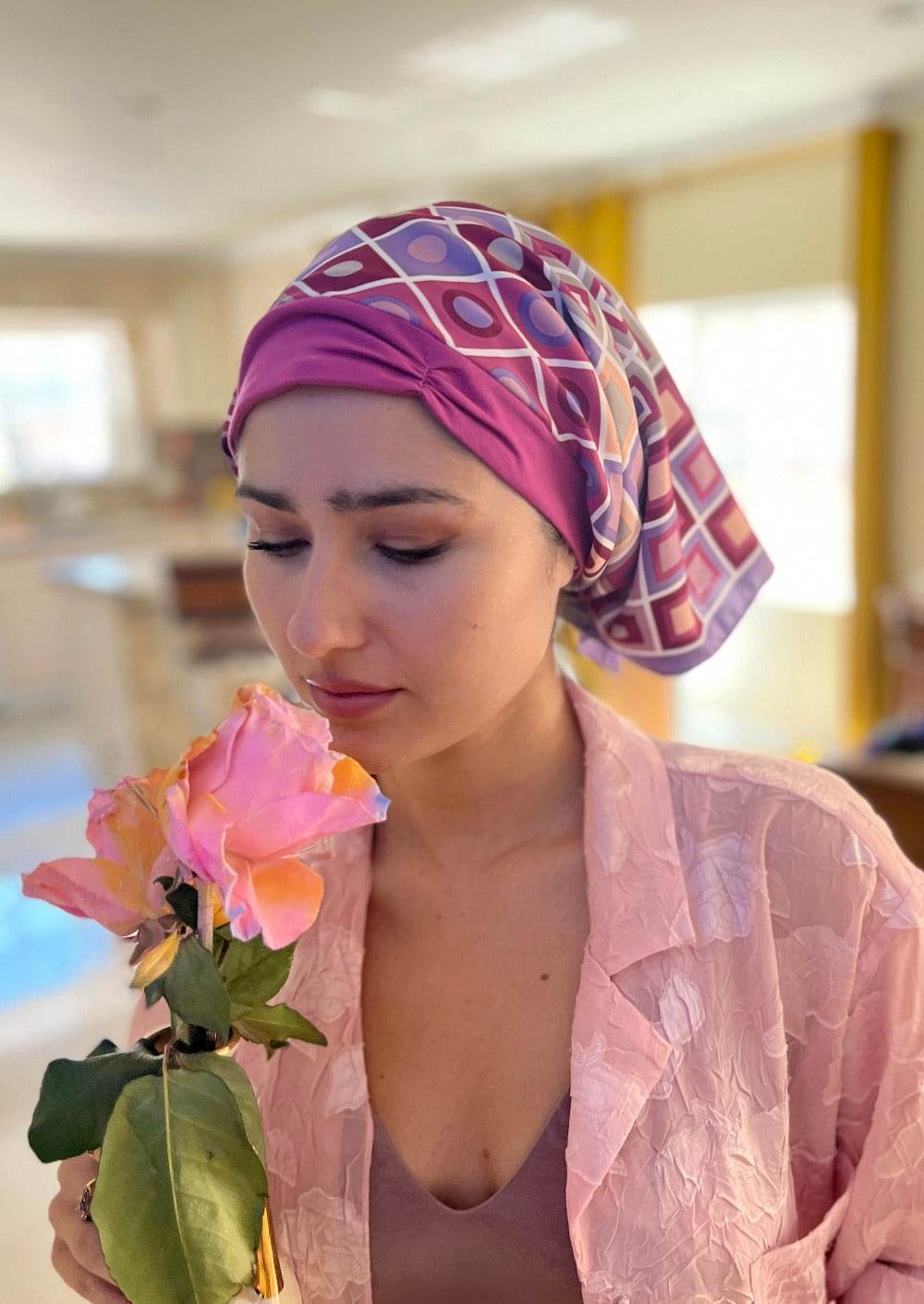 A lovely lady hold a vase with a gorgeous pink and yellow tone rose, she is smelling the flower. she has on a pink button down pj top. Her head is covered with a very attractive headscarf. The headscarf is made up of multipal layers, the base layer is a r