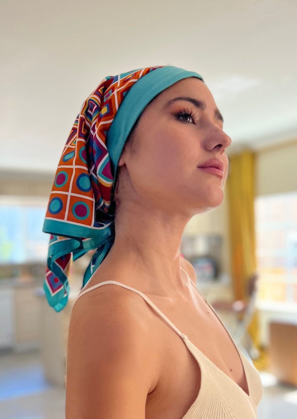 A beautiful woman, wearing an asymmetrical black shirt, stands looking to the side, in her living room. She wears a headscarf on her head, the headscarf is made up of multiple layers, the base layer is a rich teal color stretch cotton, that appears like 